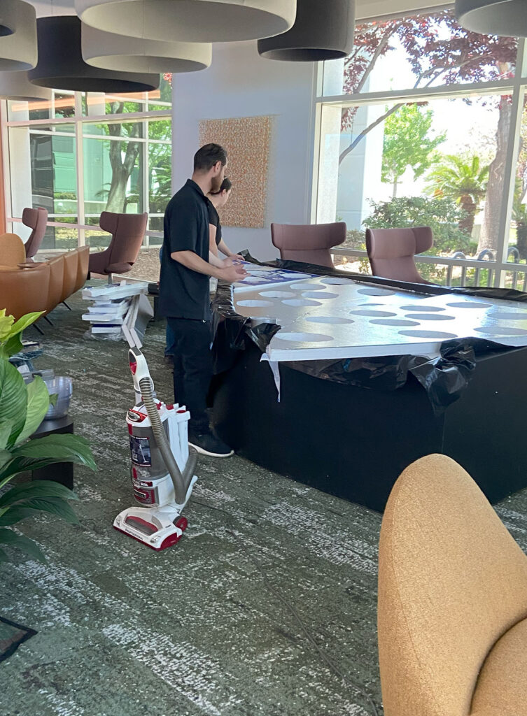 Two people work with tools on a large table. A vacuum cleaner stands nearby in a modern, well-lit room.