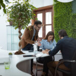 Three people collaborate at a desk in an office with plants and a large window.