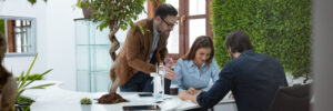 Three people collaborate at a desk in an office with plants and a large window.